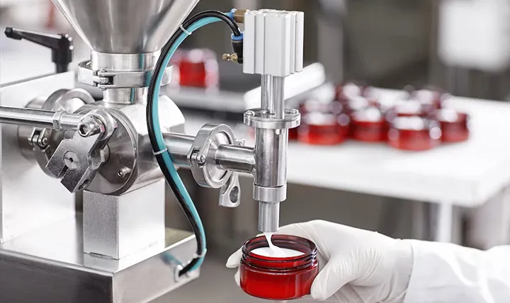 A machine filling a red cosmetic jar with white cream, held by a gloved hand.
