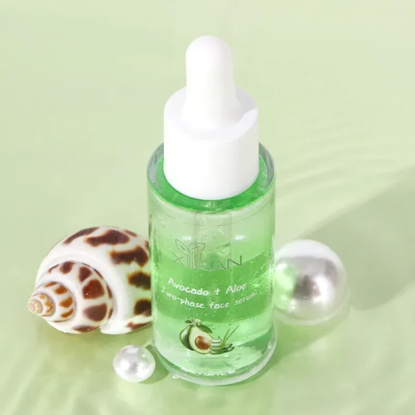 A bottle of Xiran Avocado + Aloe two-phase face serum with a seashell and pearl decoration on a light green surface.
