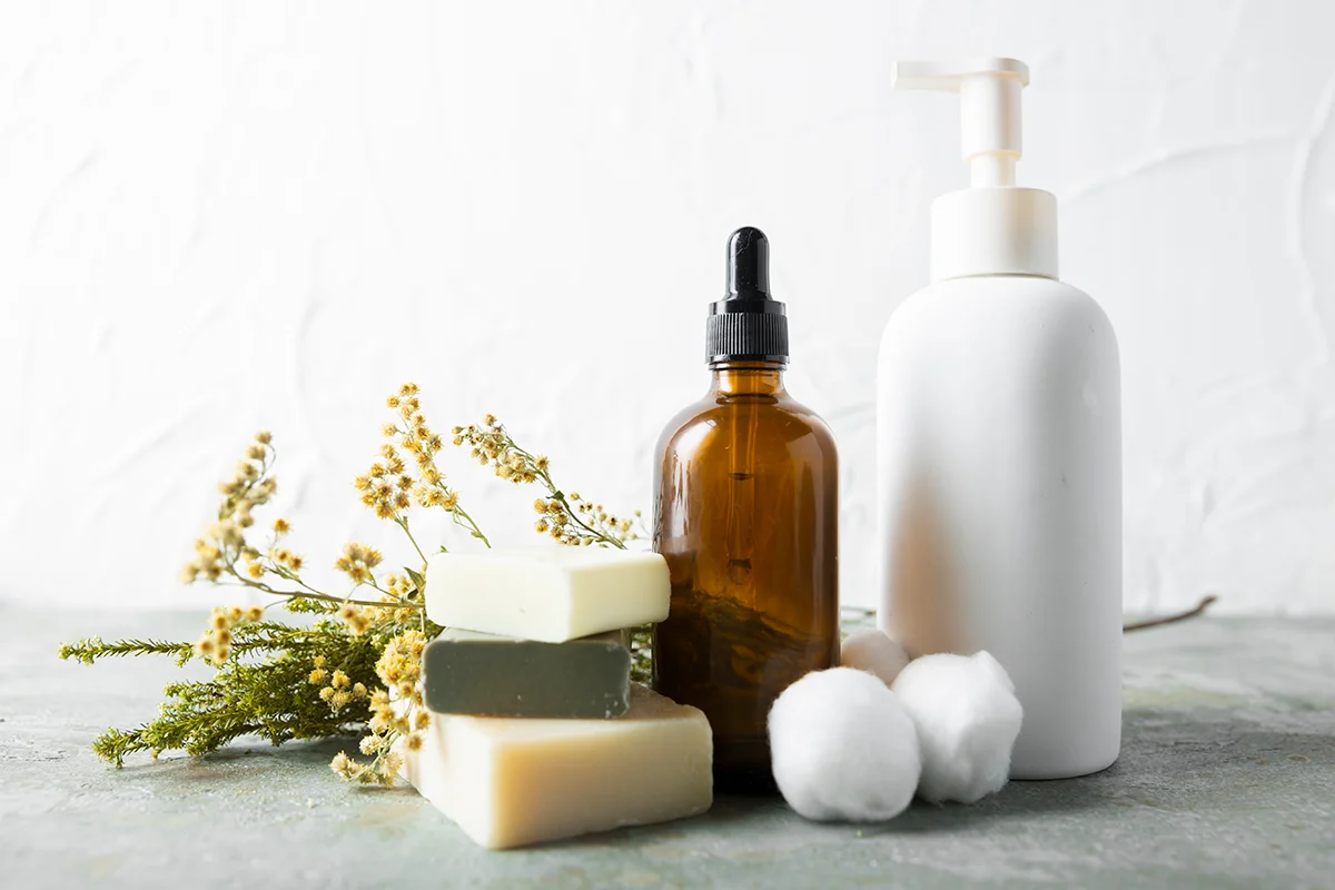 Natural skincare items including bar soap, amber bottle, and white dispenser alongside yellow flowers and cotton balls.