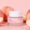 Jar of day cream labeled XIRAN with 70% peach extract and 5% niacinamide, surrounded by peaches on a pink background.