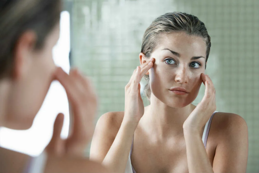 Woman wearing a white tank top looks at her reflection in a bathroom mirror with a tiled background.
