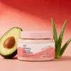 Xiran exfoliating body scrub jar with avocado half and aloe plant on a gradient pink background.