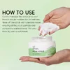Hand holding Xiran facial scrub, with cream scoop and usage instructions in background on applying the scrub.