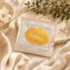 Gold collagen lip mask in glossy packaging placed on a soft, beige surface with floral decor and luxurious fabric.