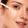 Close-up of a woman's face with liquid serum applied under her eye using a dropper. Glowing skin on a peach background.