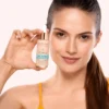 A woman wearing a yellow tank top holds a Xiran eye serum bottle with 5% caffeine and hyaluronic acid, against a neutral background.