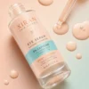 Close-up of XIRAN eye serum bottle with caffeine, hyaluronic acid, and vitamin C. Serum drops scattered on a pastel background.