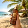 Close-up of a person's hand holding a bottle of Beach Glow suntan body oil against a sunny tropical beach background with palm trees and a hut.