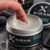 Close-up of a person dipping a finger into a jar of Xiran men's grooming pomade with firm hold, displayed on a dark marble surface with a comb and towel nearby.