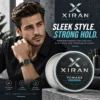 Man with styled hair wearing a black shirt and watch, seated next to a tin of Xiran men's grooming pomade for firm hold.
