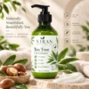 Green bottle of Xiran tea tree conditioner with pump top, surrounded by argan nuts, leaves, and conditioner cream on a natural stone surface. Product details and benefits are displayed in the background.